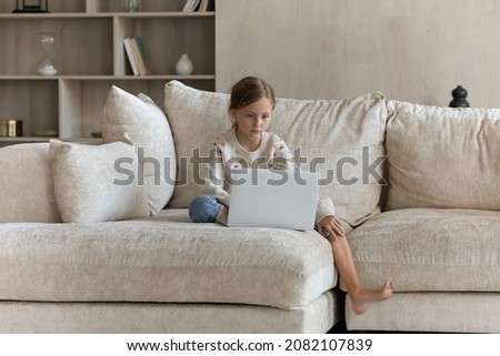 Full length focused small 7s child girl learning using computer applications alone at home, sitting on huge comfortable couch, enjoying web surfing information, studying online, modern tech addiction.