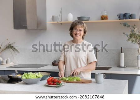 Happy senior 60s lady chopping fresh vegetables for salad at kitchen table, looking at camera, smiling. Cheerful homeowner woman, food blogger preparing ingredients for dinner. Head shot portrait