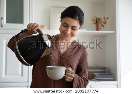 Smiling young indian ethnicity woman pouring hot water from kettle into cup, brewing tea or coffee, drinking beverage in modern kitchen, enjoying peaceful morning evening stress free carefree time. Royalty-Free Stock Photo #2082107974