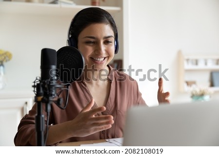 Smiling young indian female radio program host or blogger in headphones recording audio or voice acting using professional stand microphone, communicating in social network or streaming online.