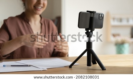 Focus on cellphone standing on table on tripod stabilizer, motivated indian young female blogger recording video or stories on smartphone web camera, talking streaming online, videoblog concept. Royalty-Free Stock Photo #2082108031