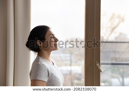 Side view calm happy inspired beautiful young indian ethnicity woman standing near window, breathing fresh air, meditating with closed eyes, imagining good future, enjoying carefree leisure time. Royalty-Free Stock Photo #2082108070
