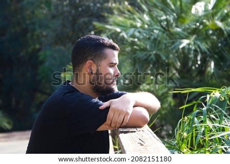 A gay man of Latin origin, dark and bearded, stands pensively looking nowhere in the park. He is sad, dazed and depressed. Concept of expressions of sadness and depression. Royalty-Free Stock Photo #2082151180
