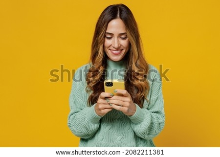 Young smiling woman 30s wearing green knitted sweater hold in hand use mobile cell phone chatting browsing internet isolated on plain yellow color background studio portrait. People lifestyle concept Royalty-Free Stock Photo #2082211381