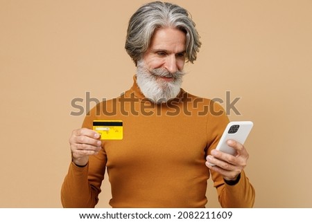Elderly gray-haired mustache bearded man in mustard yellow turtleneck shirt using mobile cell phone hold credit bank card shopping online booking tour isolated on plain pastel beige background studio