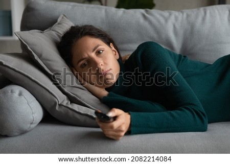 After hard working day. Exhausted millennial latin woman lying on soft couch watching evening television show news broadcast. Fatigue young hispanic female relax on cozy sofa hold tv remote in hand Royalty-Free Stock Photo #2082214084