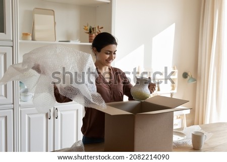 Happy young indian ethnicity woman unpacking huge cardboard box from internet store, feeling excited of receiving vase, enjoying improving interior, purchasing decorations and ware for new home. Royalty-Free Stock Photo #2082214090