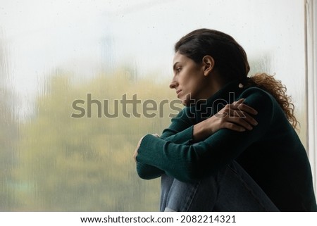 Raindrops like tears. Sad lonely latina female sit on windowsill in melancholic mood watch rain outside feeling her heart broken. Pensive young woman hug herself suffer of unanswered love. Copy space Royalty-Free Stock Photo #2082214321