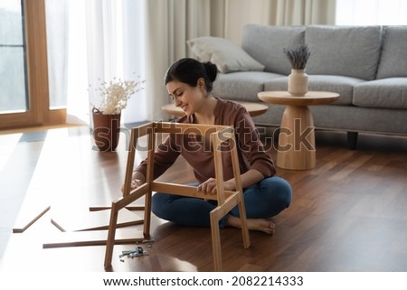 Full length smiling millennial generation indian woman sitting on warm heated floor, constructing wooden furniture in modern living room, fixing broken chair or table, improving interior at home. Royalty-Free Stock Photo #2082214333