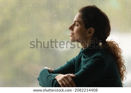 Music of rain. Side shot of serene latin woman relax by window with closed eyes listen sound of raindrops feel pleasure. Calm young lady enjoy breathing fresh ozonized air after rainstorm. Copy space. Royalty-Free Stock Photo #2082214408
