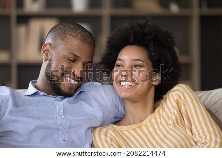 Happy African American couple relaxing at home, talking and laughing, sitting close, hugging. Young husband and wife joking and having fun, embracing on sofa, enjoying being dogether