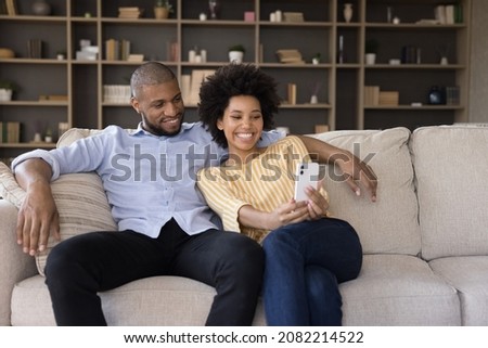 Cheerful millennial Black couple using mobile phone, taking selfie, resting on sofa at home, embracing, smiling at smartphone screen. Happy guy and girl in love with cellphone making video call