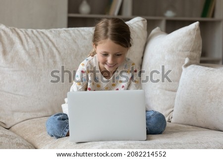 Full length addicted to modern tech joyful little cute kid girl sitting on couch with computer, enjoying playing entertaining online game, communicating in social network, watching funny video content