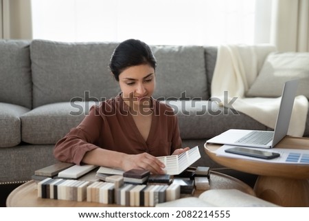 Happy motivated beautiful young indian woman sitting at table with different swatches, choosing materials planning own apartment redecoration improvement or creating interior design alone at home.