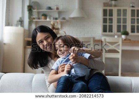 Cheerful Asian babysitter tickling little cute boy. Preschool 6s son enjoy playtime with loving young mother, family sit on sofa play together at home. Custody, happy motherhood, leisure fun concept Royalty-Free Stock Photo #2082214573