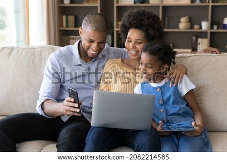 Happy African American parents and daughter kid resting on couch together, enjoying leisure with digital devices, holding, sharing laptop, smartphone, tablet, laughing, watching online content Royalty-Free Stock Photo #2082214585