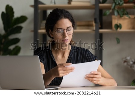 Important papers. Serious hispanic female manager in glasses engaged in office paperwork study project documentation cover letter. Young woman hr reading resume thinking on applicant cv at workplace Royalty-Free Stock Photo #2082214621