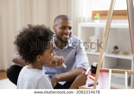 Happy cute Black little kid and dad drawing on chalkboard at home, using colorful chalks and whiteboard, laughing, having fun, enjoying leisure, playtime, entertainment, craft creative hobby