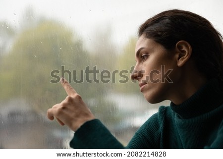 Crying rain. Thoughtful millennial hispanic woman suffer from depression sit by large window running finger over glass watching raindrops. Pensive unhappy young lady lost in sad thoughts. Copy space