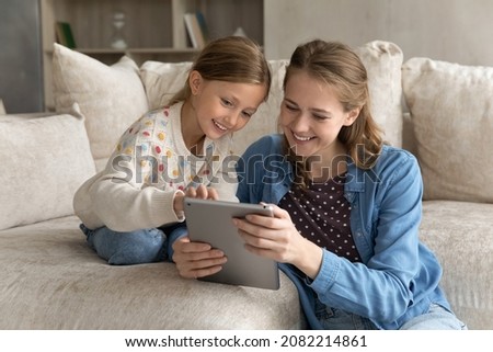 Happy pretty adorable child girl showing funny apps on digital tablet to caring young mother, choosing goods shopping in internet store, relaxing together on cozy sofa, modern tech addiction concept.
