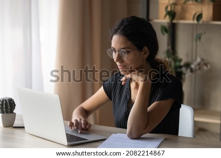 Interesting task. Focused young hispanic woman in eyewear sit by desk at home work online by laptop pc. Millennial female office worker employee search information at corporate network read e document Royalty-Free Stock Photo #2082214867