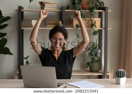 Euphoric mood. Excited young latin lady worker sit by desk raise hands up after successful finish hard work on business project. Female manager celebrate triumph of making great deal by office laptop Royalty-Free Stock Photo #2082215041