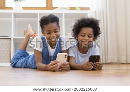 Sweet African sibling children relaxing on warm floor, enjoying leisure time with digital gadgets, using learning application on mobile phones, chatting on social media, watching online media content Royalty-Free Stock Photo #2082215089