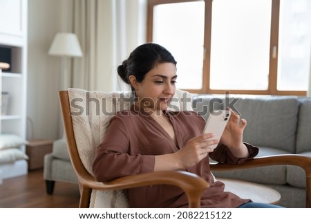 Relaxed young beautiful indian woman using cellphone software applications, sitting in comfortable armchair, playing mobile games, shopping in internet store, web surfing or communicating distantly. Royalty-Free Stock Photo #2082215122