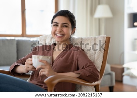 Portrait of smiling beautiful indian ethnicity woman resting in cozy armchair with cup of coffee or tea in hands, enjoying carefree lazy relaxed morning weekend time lone in modern living room. Royalty-Free Stock Photo #2082215137