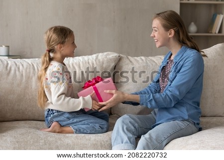 Cheerful young mother giving wrapped gift box to excited small kid daughter, congratulating with happy birthday or making surprise present, sitting together on comfortable sofa in living room. Royalty-Free Stock Photo #2082220732