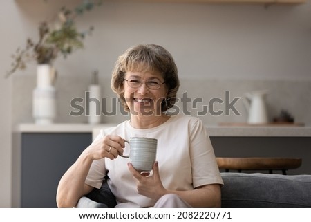 Happy older woman in glasses enjoying good morning, drinking hot coffee at home, relaxing on comfortable sofa in cozy living room, holding ceramic mug, cup of tea, smiling. Head shot portrait