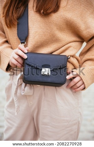 Closeup of black small handbag in woman's hand. Fall spring fashion outfit camel sweater and trendy white jeans. Fashion detail, stylish small bag.   Royalty-Free Stock Photo #2082270928