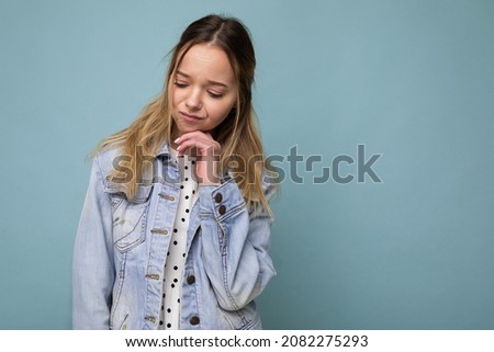 Photo of young sad upset beautiful blonde woman with sincere emotions wearing denim blue jacket isolated over blue background with copy space