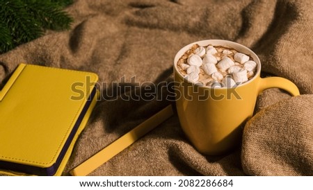 Yellow mug with drink and marshmallows with hard shadow. Yellow notepad and pen lie next to it. Tree branch is visible on left. Place for your text. Selective focus.