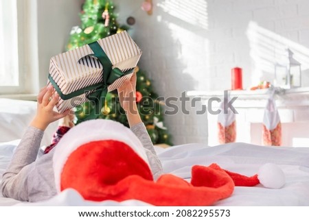 Little boy in a red Santa hat and pajamas holds a gift box in his hands while lying in a bed in front of a Christmas tree at home at morning. The child looks at the present, back view.