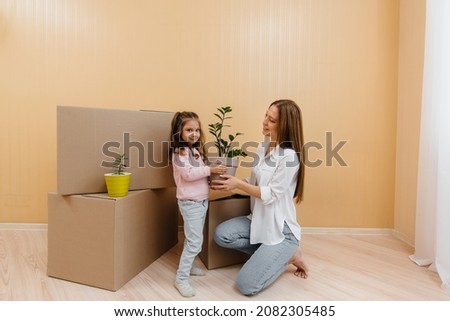 A young woman stands with a houseplant next to her little daughter after moving into a new apartment against the background of cardboard boxes. Purchase of real estate. Housewarming, delivery.