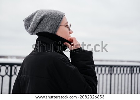 Pensive thoughtful young woman with short hair in hat, eyeglasses, black coat walking on a winter city street and looking away. Enjoying weather and having fun. Candid people