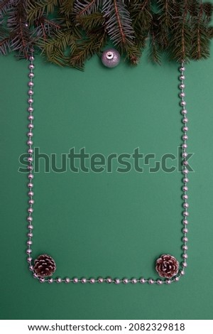 Evergreen twigs with one silver Christmas decoration and cones on a green background. Copy space framed with silver pearls. Minimalistic flat lay concept.