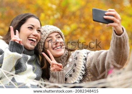 Two happy friends taking selfie with smart phone on hammock in autumn