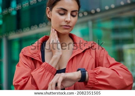Cropped shot of serious woman checks pulse on neck monitors fitness activity has quick heart rate wears red jacket poses outdoors against blurred background controls her health. Devices for sport Royalty-Free Stock Photo #2082351505