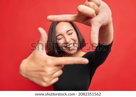 Beautiful cheerful woman makes frame gesture smiles positively keeps eyes closed composes picture idea dressed in black jumper poses against vivid red background gazes at camera through hands