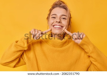 Portrait of cheerful young woman smiles broadly indicates index fingers at face wears casual jumper expresses positive emotions isolated over vivid yellow background. Look at my brilliant smile Royalty-Free Stock Photo #2082351790