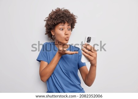 Lovely positive young woman sends air kiss at smartphone camera keeps lips folded expresses love to boyfriend on distance wears casual blue t shirt isolated over white background blows mwah.