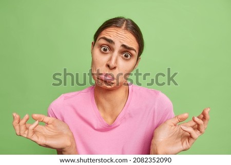 Hesitant clueless unaware young woman spreads palms looks with sad doubtful expression purses lower lip poses against bright green background. Discontent questioned female model shrugs shoulders