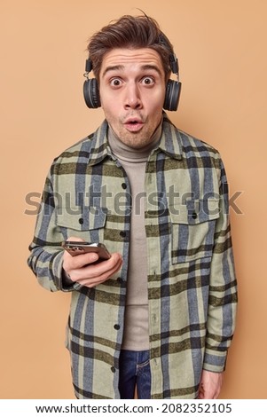 Photo of emotional shocked young guy stares impressed at camera has unbelievable gaze holds mobile phone listens music via headphones wears casual checkered shirt isolated over beige background