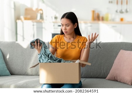 Dissatisfied young asian woman opening box from online store and looking at wrong delivered item, unhappy with received shirt, disliking clothing after unboxing package Royalty-Free Stock Photo #2082364591