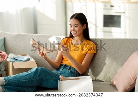 Excited asian lady unpacking parcel, making photo of her new shoes on smartphone for positive online store feedback, sitting on sofa, copy space. Positive lady satisfied with service