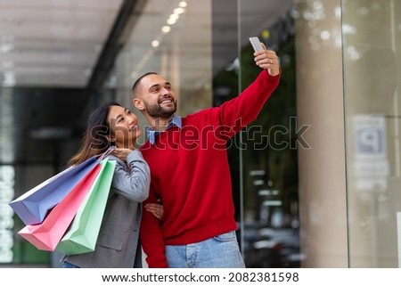Young international couple with bright paper bags taking selfie near big shopping centre, copy space. Millennial multiracial spouses photographing themselves on street next to city mall