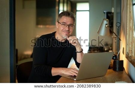 Older man sitting at desk in dark cosy room working on laptop computer in home office. Mature age, middle age, mid adult casual man in 50s, confident happy smiling. Royalty-Free Stock Photo #2082389830