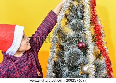 A joyful boy wearing a Santa Claus hat decorates the Christmas tree with New Year's toys and glittering tinsel.